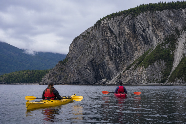 Two kayakers paddling on calm water in Norris Point with dramatic cliffs and forested mountains in Gros Morne National Park.
